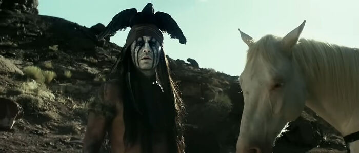 Actor in miscast role wearing face paint and black headpiece standing next to a white horse in a rocky outdoor setting