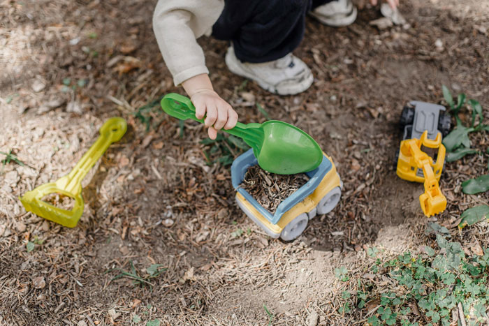Child playing with toys, digging in dirt and causing damage to neighbor lawn with green shovel and plastic truck.