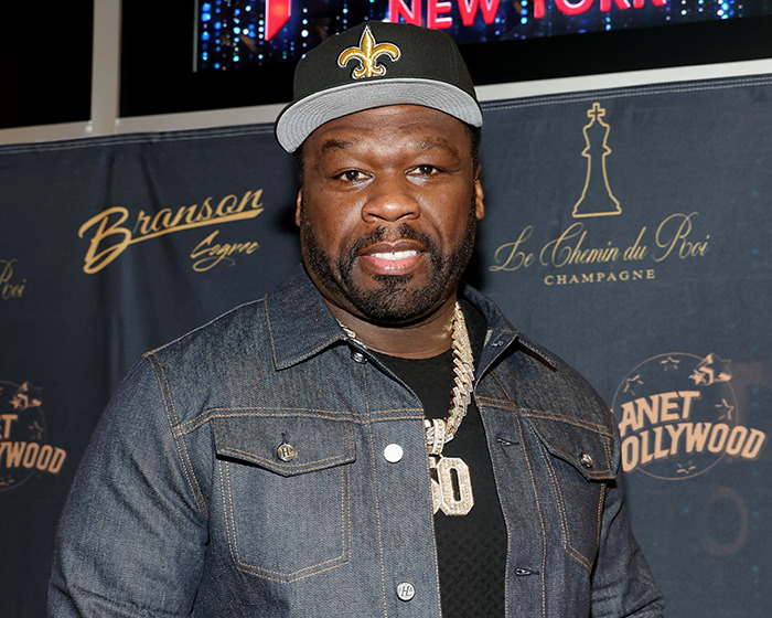 50 Cent posing at event wearing a denim jacket and a black cap with a gold emblem, promoting Free Diddy shirts.