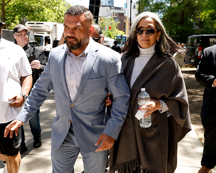 Man in light blue suit escorting woman in sunglasses and dark shawl, walking outdoors with people around in a city setting.