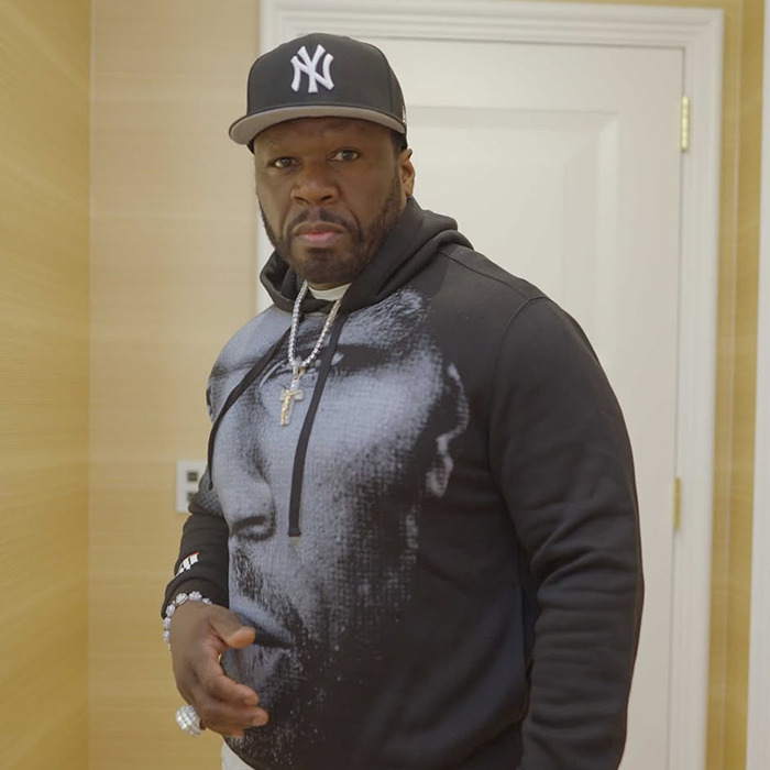 50 Cent wearing a black hoodie with a face print and a New York Yankees cap, posing indoors.