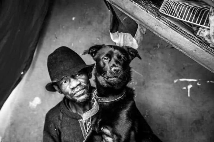 Homeless man wearing a hat holding his black dog inside a shelter, capturing lives of homeless people and their dogs.