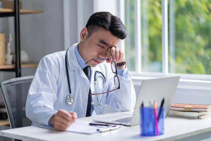 Doctor in a white coat sitting at a desk, rubbing eyes tiredly while reviewing papers on laptop, illustrating life blunders.