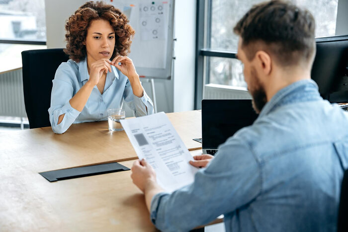 Woman nervously facing a man reviewing her resume during an interview, feeling like being picked last in adult life moments.