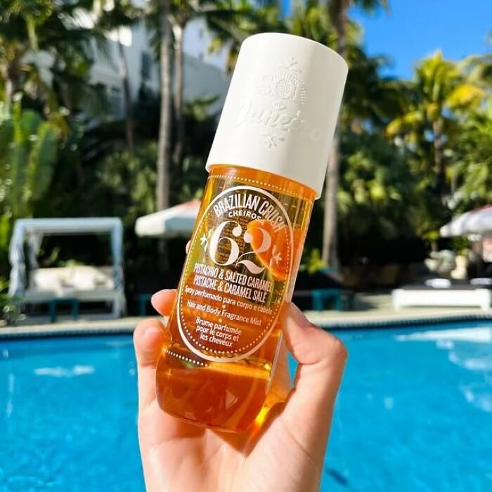 Hand holding Brazilian Crush hair and body fragrance mist by a pool with palm trees in the background for warm weather finds.