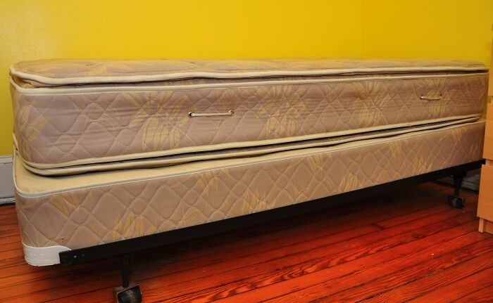 A beige mattress placed on a wheeled bed frame against a yellow wall and wooden floor, showing excessive laziness.