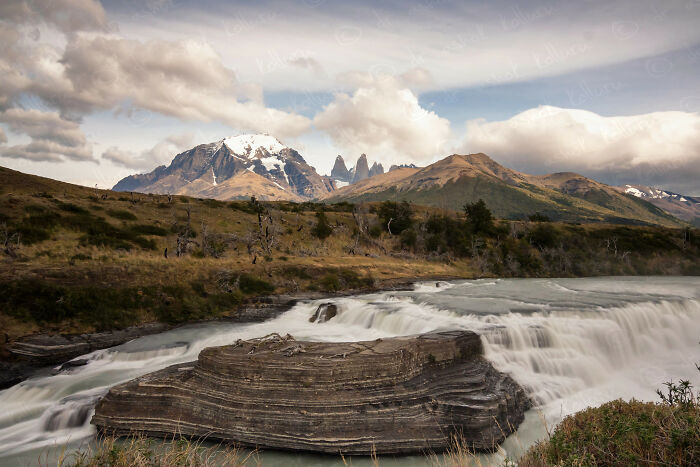 A stunning natural wonder with a waterfall, rocky formations, and mountains under a cloudy sky showing the world’s magic.