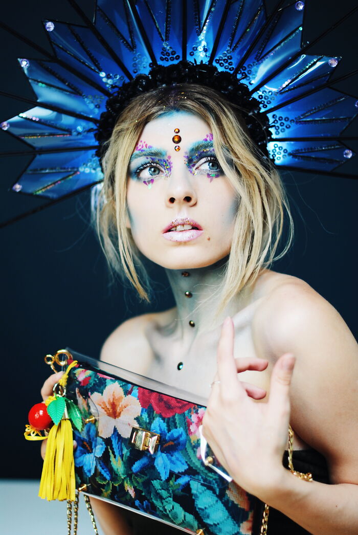 Woman with intricate makeup and jeweled face holding floral clutch, styled with bold headpiece showcasing incredible makeup looks.