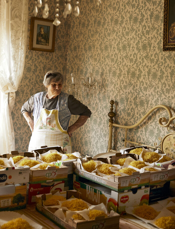 Elderly woman in a vintage kitchen surrounded by boxes of fresh pasta in a food photography setting.