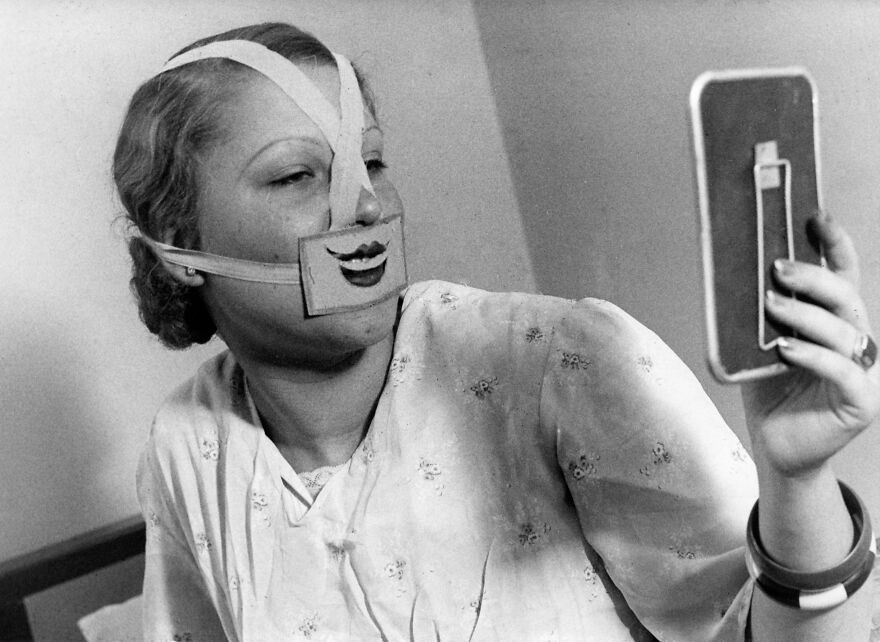 A woman in 1937 Budapest wearing a bizarre medical mask designed to fight depression, holding a mirror.