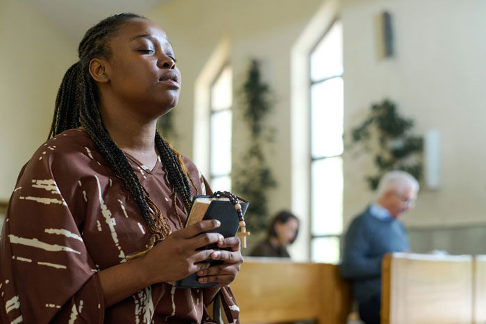 Woman holding a rosary and Bible, eyes closed in prayer inside a church, relating to people who escaped from cults.