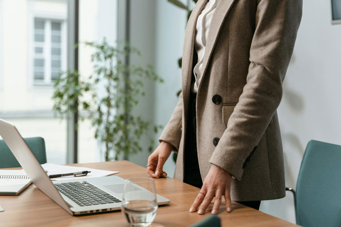 Person in charge standing by a desk with a laptop and documents, conveying authority in a professional office setting.