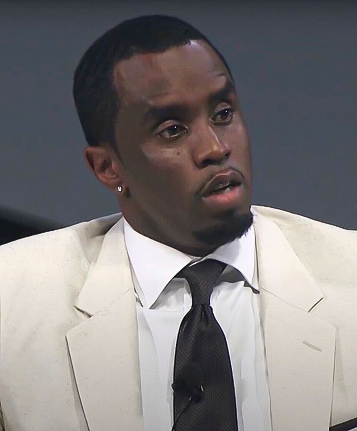 Diddy wearing a white suit and black tie, appearing serious during a discussion related to the alleged victim testimony case.