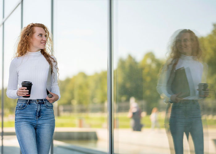 Woman holding coffee cup outside near glass wall, reflecting things everyone does but doesn’t talk about moments.