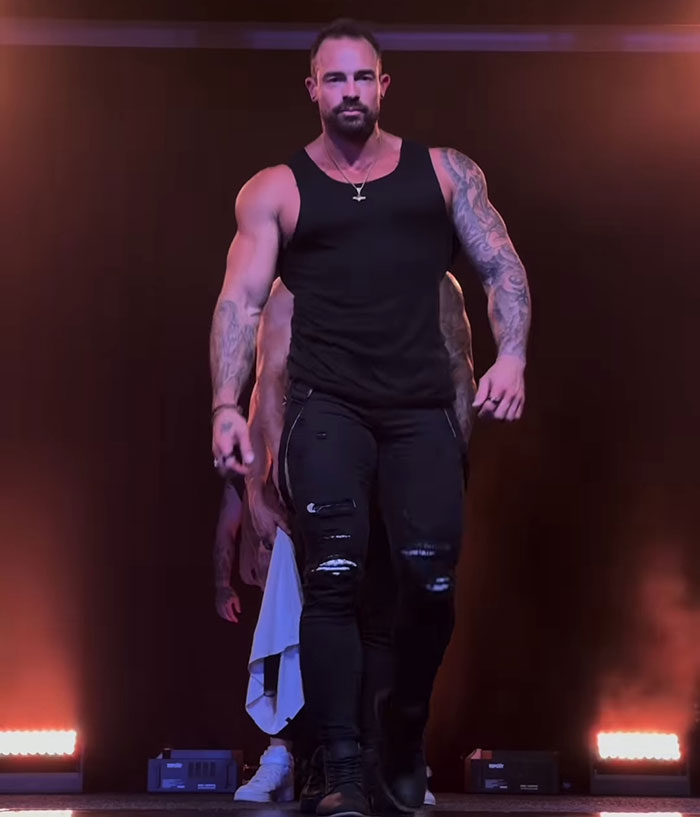 Muscular man with tattoos and black tank top walking on stage, symbolizing former pole dancer turned monk story.