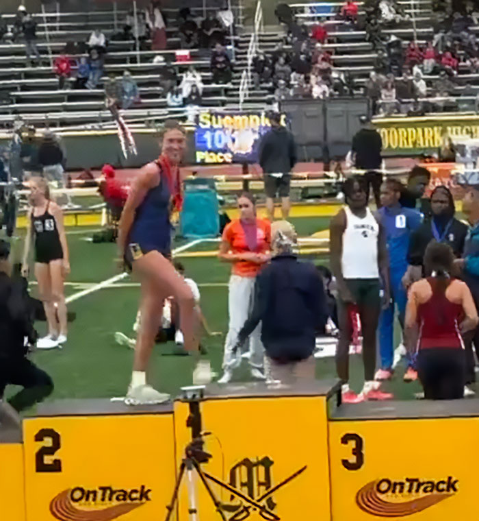 High school girl standing defiantly on first-place podium at track event after losing to trans athlete.