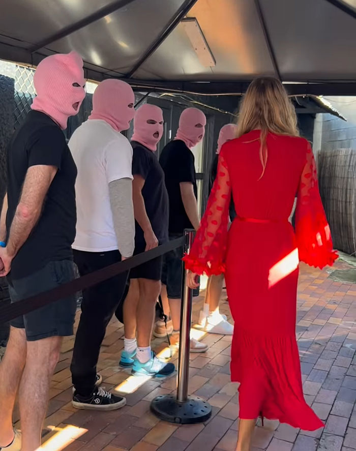Woman in red dress walking past men in pink ski masks, illustrating bleeding and hospital update for adult content creator.