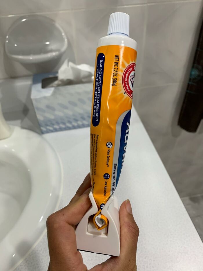 Hand holding a toothpaste tube with a white and orange cap, using a tube squeezer for everyday finds fixing annoyances.