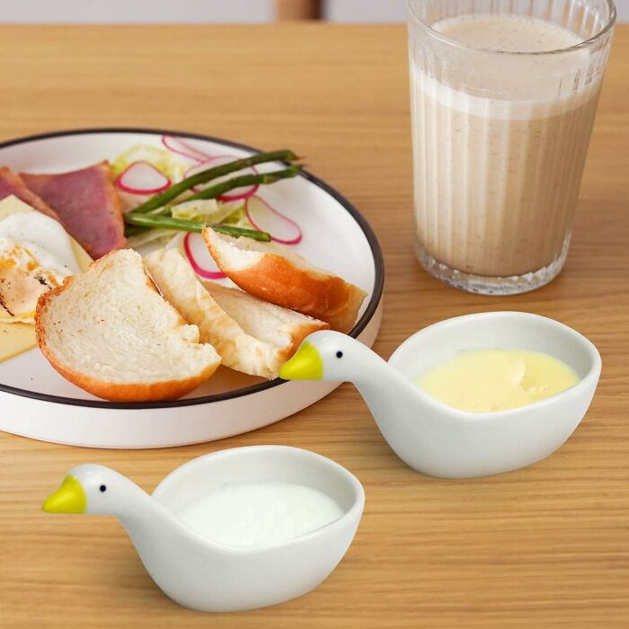 Two white ceramic goose-shaped sauce dishes next to a breakfast plate and a glass of smoothie on a wooden table.