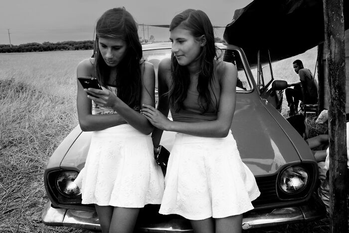 Two young women in white skirts leaning on a car, one focused on a phone, capturing the essence of humanity in black and white.