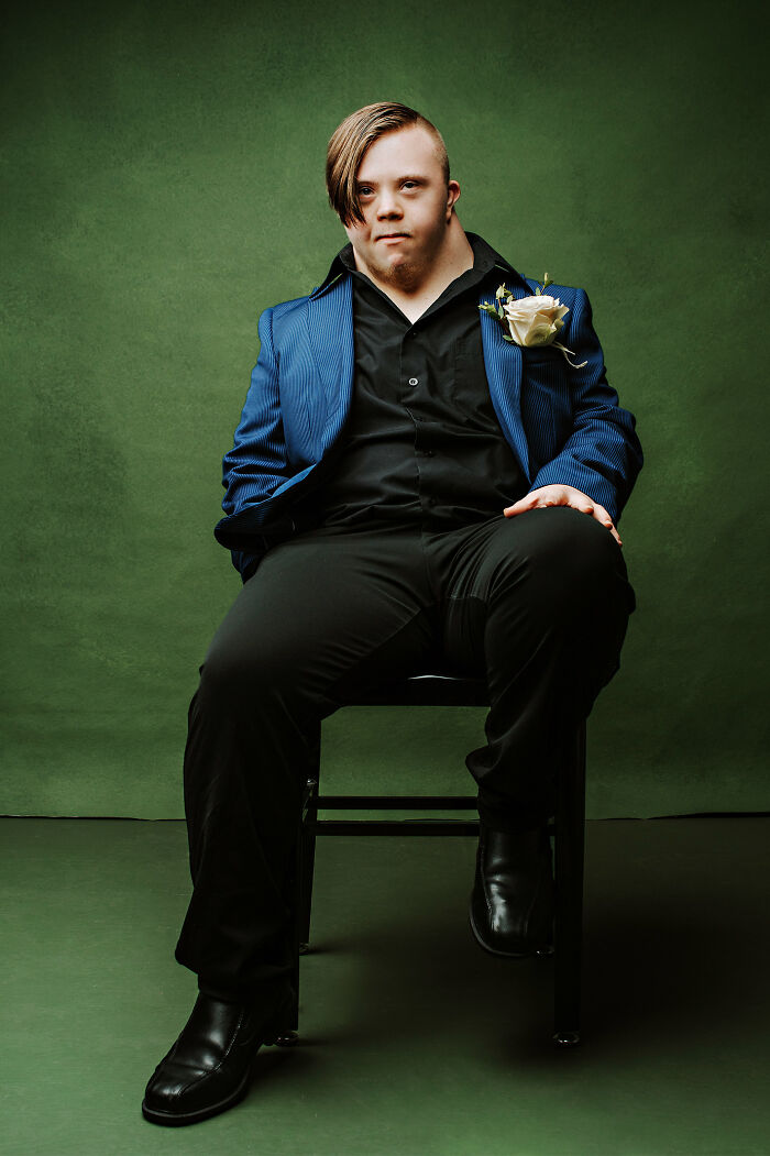 Young man with Down syndrome in a blue jacket and black outfit sitting confidently against a green backdrop promoting inclusion.
