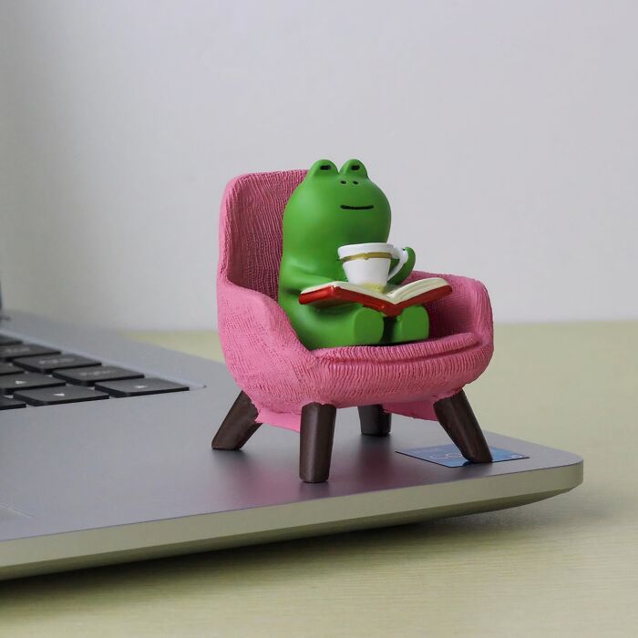 Green frog figurine holding a cup and book, sitting on a pink chair as a quirky office supply on a laptop.