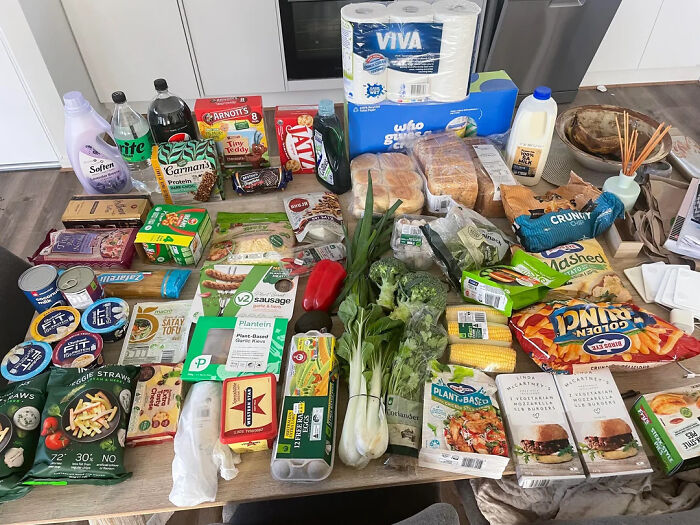 Grocery store haul with various fresh vegetables, snacks, beverages, and household essentials displayed on a kitchen table.
