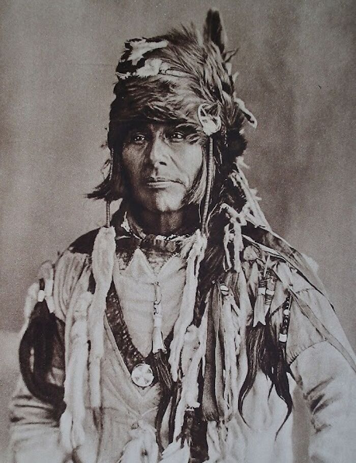Native American man in traditional attire and feathered headdress in a powerful portrait from over a century ago.
