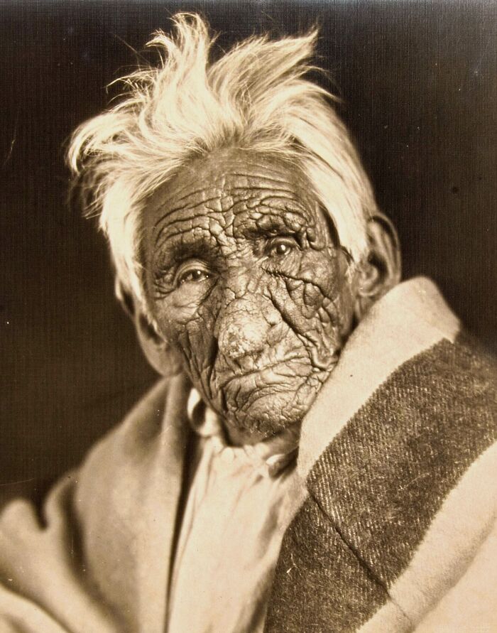 Elderly Native American man with deeply wrinkled face and white hair wrapped in a patterned blanket in a powerful portrait.