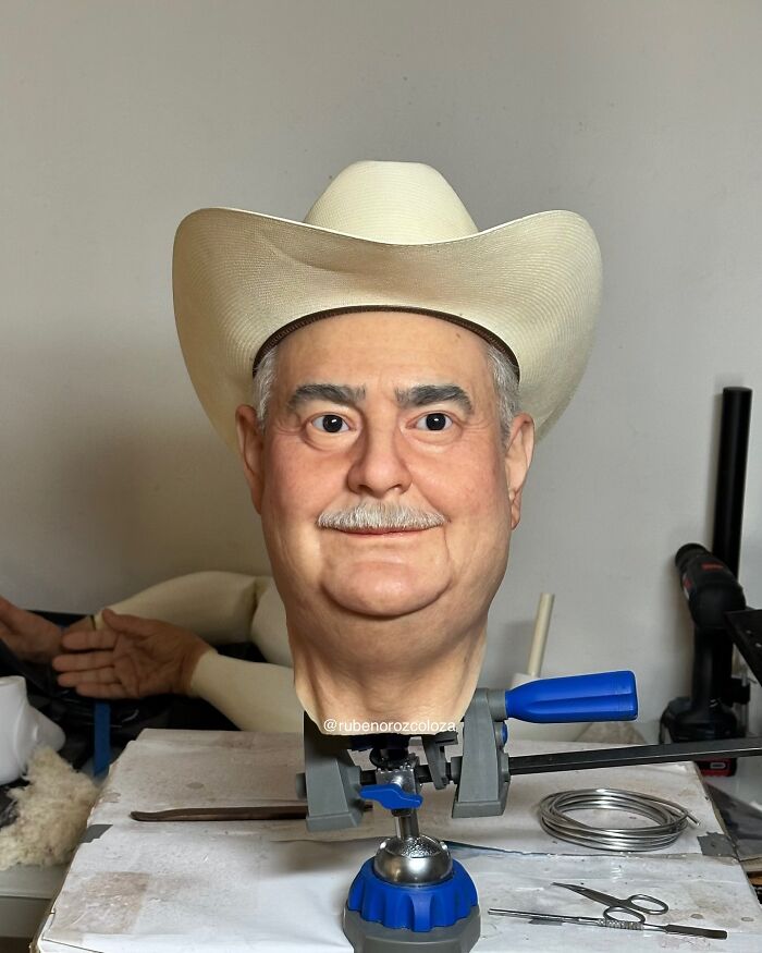Hyper-realistic sculpture of a man wearing a cowboy hat displayed in an artist's workshop by Ruben Orozco Loza.