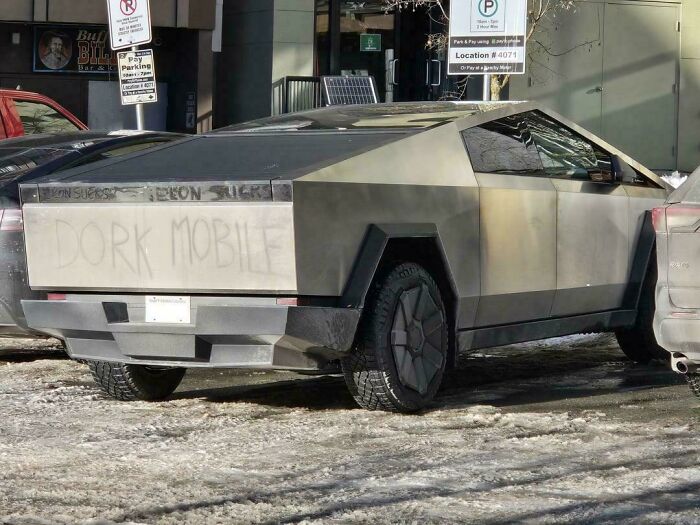 Matte gray Tesla Cybertruck parked on a snowy street with graffiti reading dork mobile on rear panel.