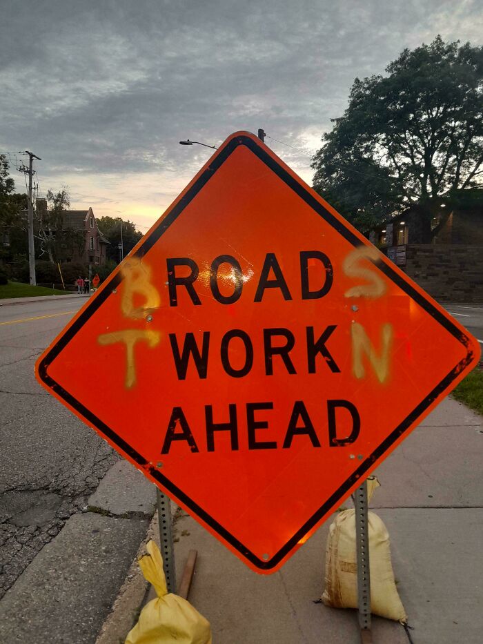 Road work ahead sign with yellow spray paint graffiti, showing wholesome vandalism brightening the street scene.