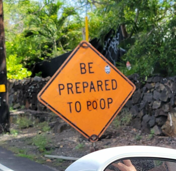 Humorous street sign altered by wholesome vandals reading be prepared to p**p on a roadside with greenery and rocks.