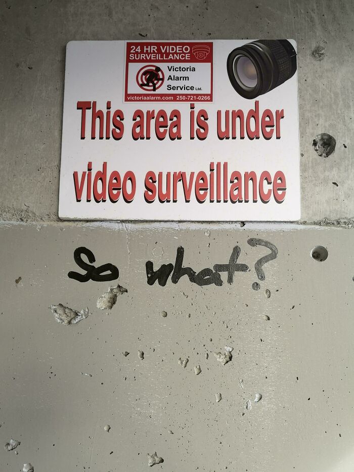 Sign warning of video surveillance with graffiti reading "so what?" on a concrete wall, showing wholesome street vandalism.