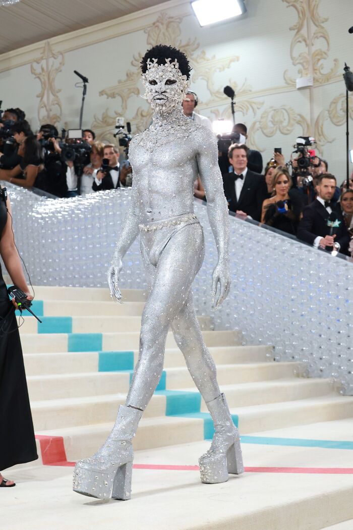 Celebrity in a sparkling silver bizarre Met Gala outfit with a bejeweled mask and platform boots on the red carpet.