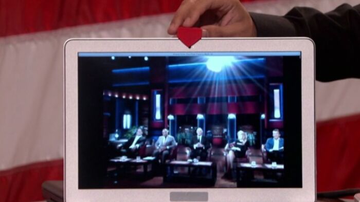 Hand holding a miniature red shark fin on a laptop showing the Shark Tank panel in a dimly lit studio setting.