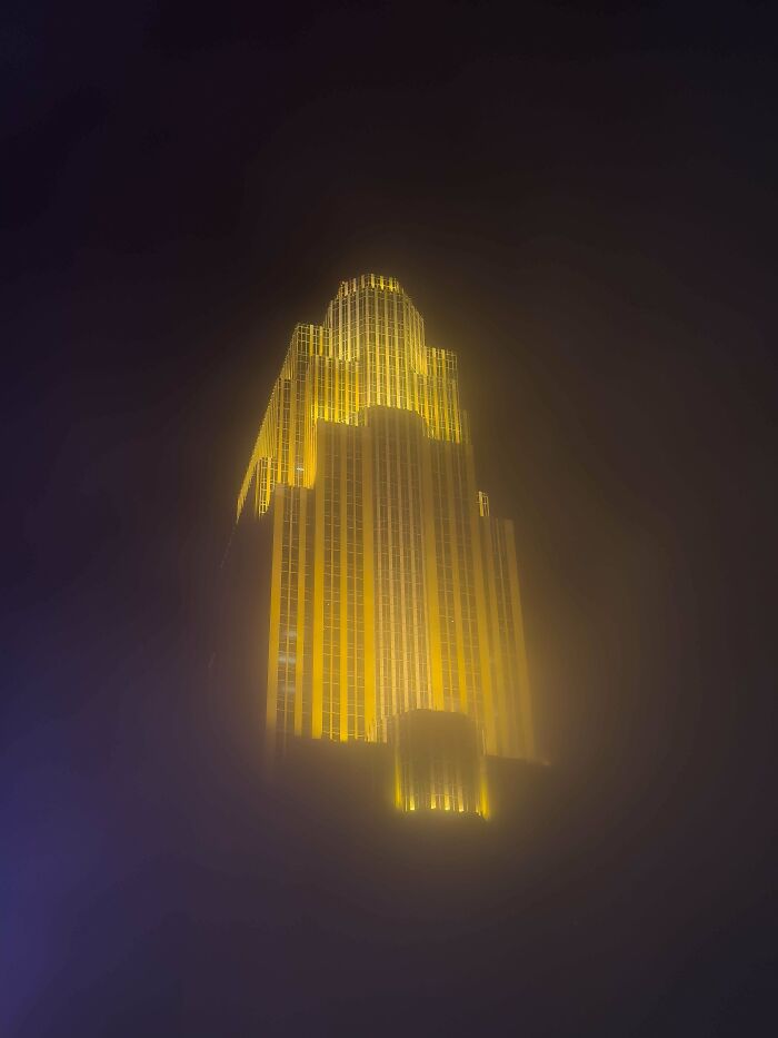 Tall illuminated skyscraper glowing yellow in thick fog, showcasing a scarily big structure linked to megalophobia.