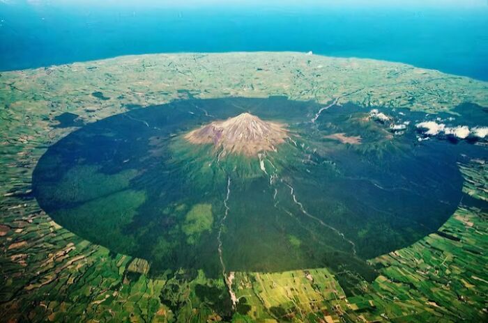 Aerial view of a scarily big volcano casting a massive shadow over surrounding fields, showing megalophobia scale.