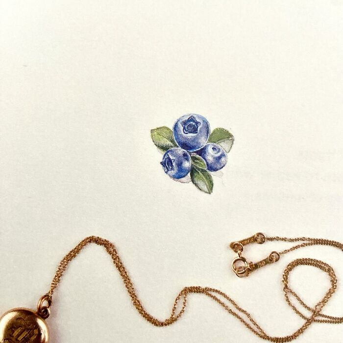 Tiny one-inch painting of blueberries on paper next to a gold chain necklace, showcasing detailed everyday art by Brooke Rothshank.