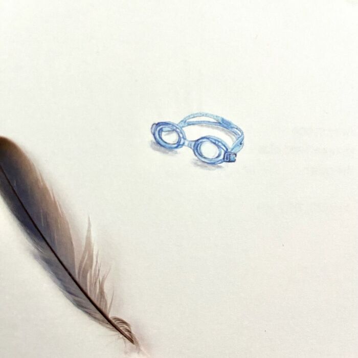 Tiny one-inch painting of blue swimming goggles next to a feather on a plain white background.