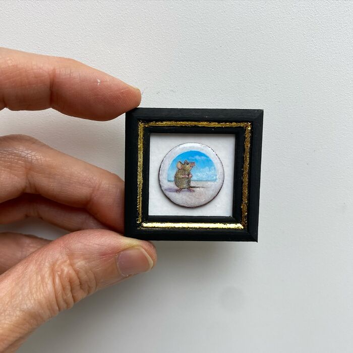 Hand holding a tiny one-inch painting of a mouse with a blue sky background in a small black and gold frame.