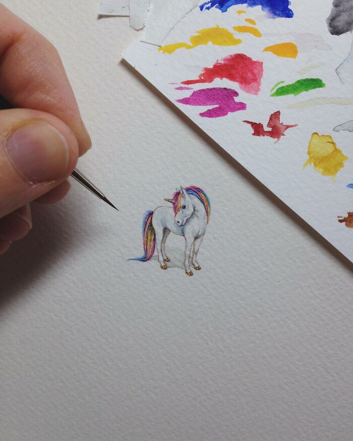 Hand painting a tiny one-inch unicorn with colorful mane on white paper, showcasing detailed miniature art by Brooke Rothshank.