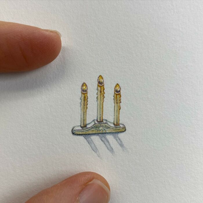 Tiny one-inch painting of three lit candles on a metal holder, shown next to fingers for scale by Brooke Rothshank.