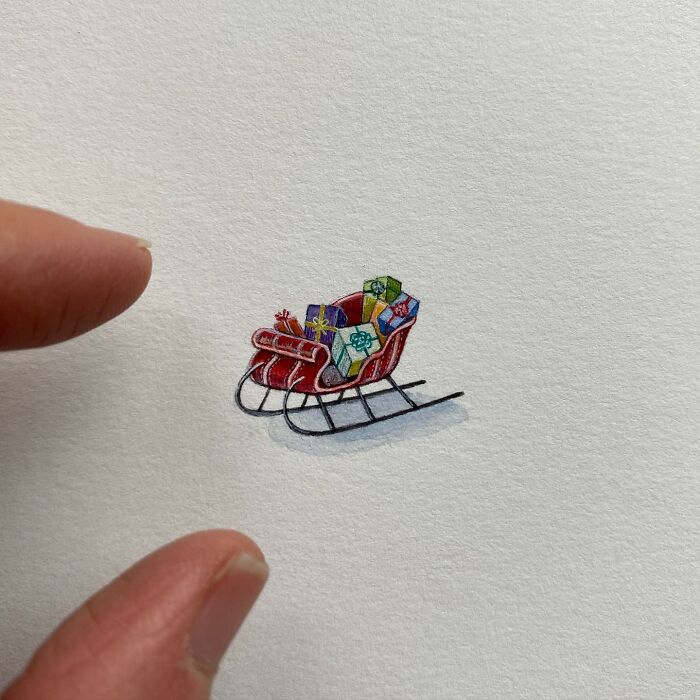 Tiny one-inch painting of a red sleigh filled with colorful gifts, shown next to fingers for scale.