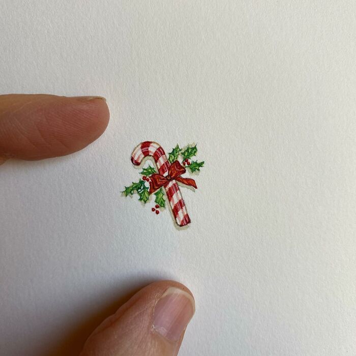 Tiny one-inch painting of a candy cane with holly, held between fingers, showcasing detailed everyday art by Brooke Rothshank.