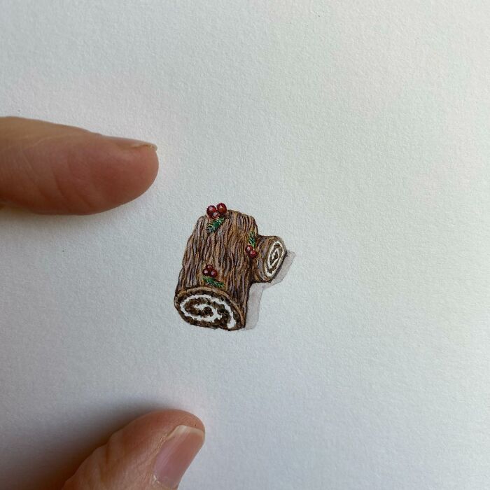 Tiny one-inch painting of a festive Yule log held between two fingers by artist Brooke Rothshank on white paper.