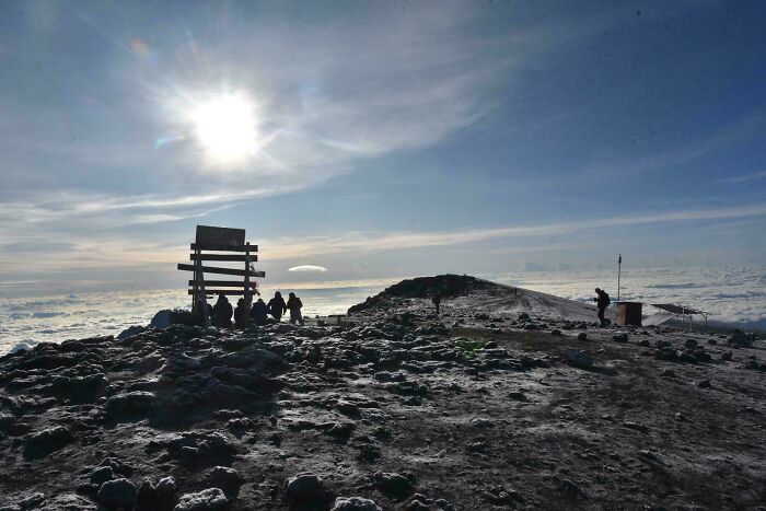 Hikers resting near a wooden sign on a rocky mountain peak under a bright sun and expansive sky, alternate angles in history.