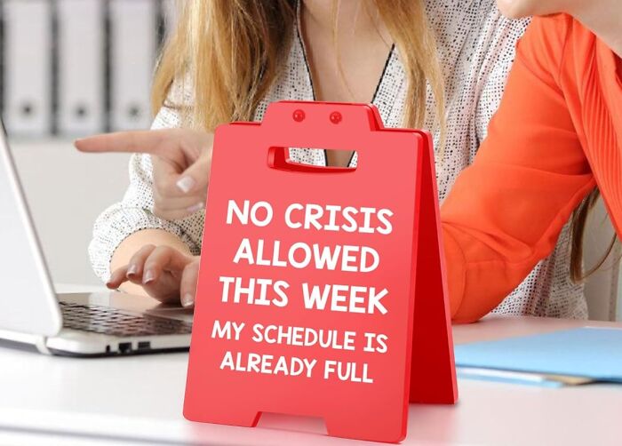 Red desk sign with office supplies saying no crises allowed this week my schedule is already full near laptop and coworkers.