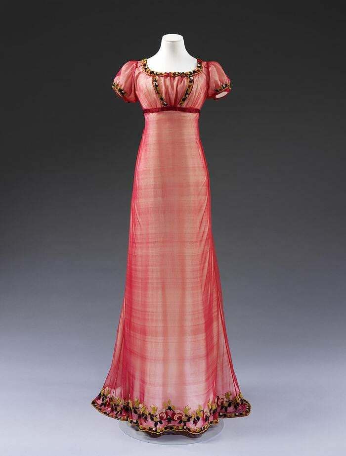 Empire waist pink vintage dress with floral trim, showcasing centuries of fashion evolution on a mannequin against gray background.