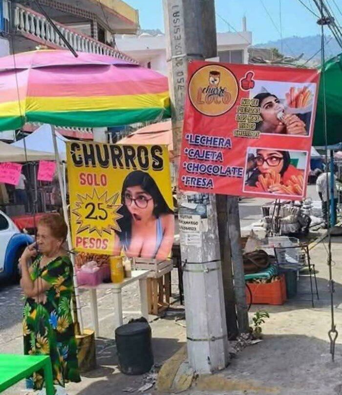 Street vendor signs for churros at 25 pesos, featuring surprising images, creating a blursed image effect in a busy market scene.